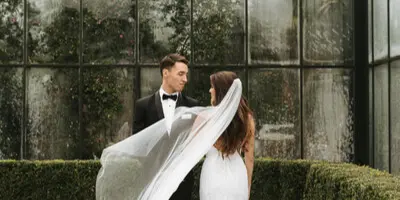 Bride and groom in wedding attire standing in front of a glass wall with greenery