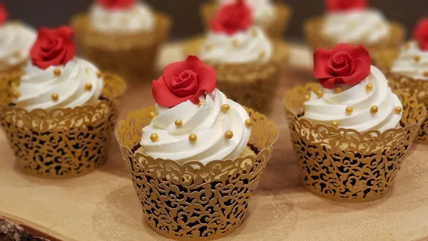 A group of cupcakes with white frosting, gold sprinkles, and red rose toppers in gold lace wrappers.