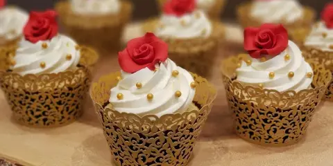 A group of cupcakes with white frosting, gold sprinkles, and red rose toppers in gold lace wrappers.