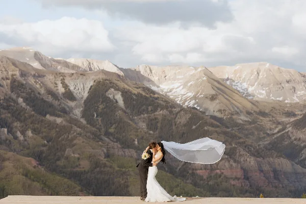 A bride and groom kissing on a wooden platform with a mountain range in the background.