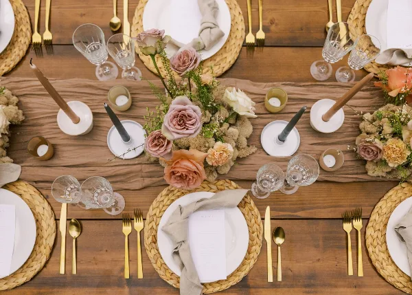 Overhead view of a rustic wooden wedding table setting featuring woven placemats, gold flatware, and a floral centerpiece with dusty pink roses and candles.