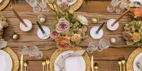 Overhead view of a rustic wooden wedding table setting featuring woven placemats, gold flatware, and a floral centerpiece with dusty pink roses and candles.