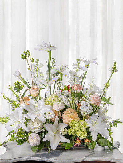 A large, elegant floral arrangement featuring white lilies, roses, and greenery on a silver tray.