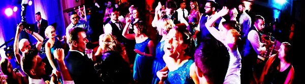 A crowded dance floor at a wedding reception with guests dancing under blue and purple lighting.