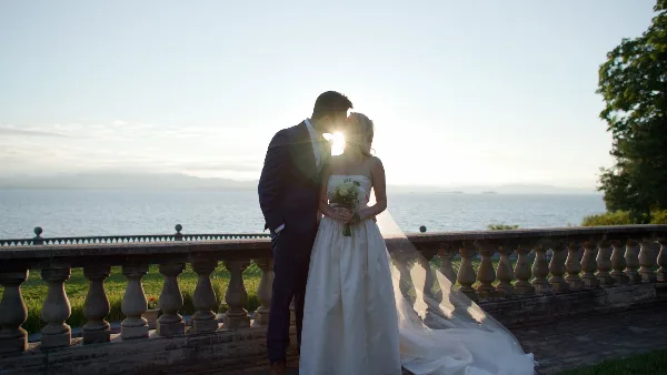 A bride and groom standing together on a stone balcony overlooking a body of water at sunset.