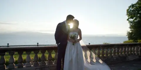 A bride and groom standing together on a stone balcony overlooking a body of water at sunset.