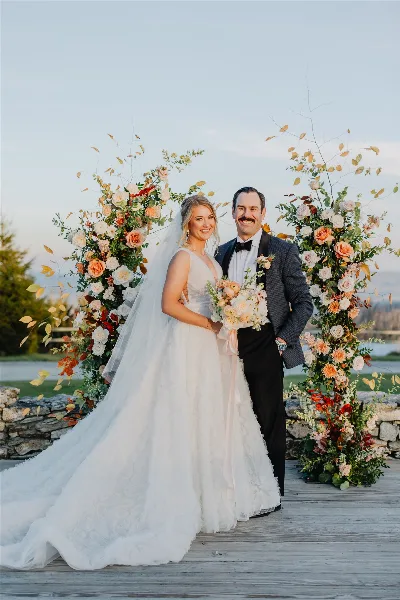 A bride and groom standing together in front of a large floral archway at a wedding venue.