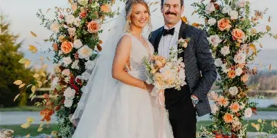 A bride and groom standing together in front of a large floral archway at a wedding venue.