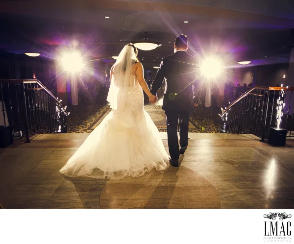 A bride and groom walking down a staircase in a ballroom, viewed from behind.