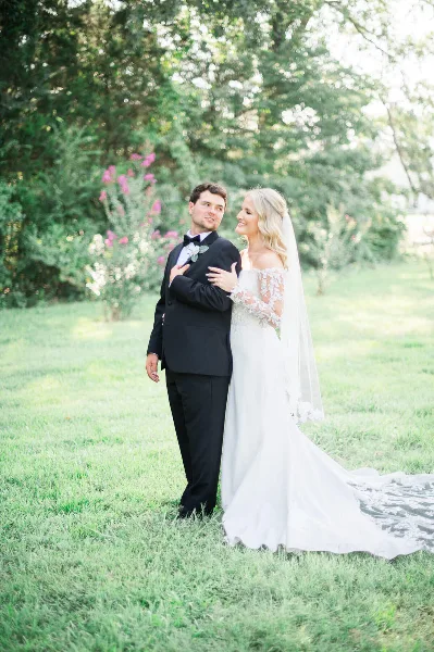 A bride and groom standing together in a grassy field with trees in the background.
