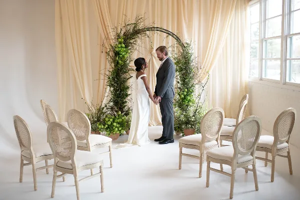 A wedding couple stands beneath a large floral archway with greenery and white flowers, surrounded by chairs in a bright room with cream drapery.