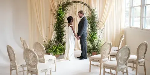 A wedding couple stands beneath a large floral archway with greenery and white flowers, surrounded by chairs in a bright room with cream drapery.