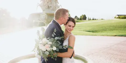 A bride and groom embracing in front of a stone fountain during a wedding.