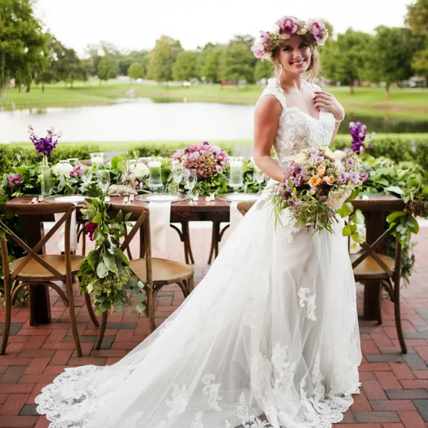 A bride in a white lace gown holds a large floral bouquet and wears a floral crown, standing in front of a wedding reception table decorated with flowers.