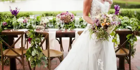 A bride in a white lace gown holds a large floral bouquet and wears a floral crown, standing in front of a wedding reception table decorated with flowers.