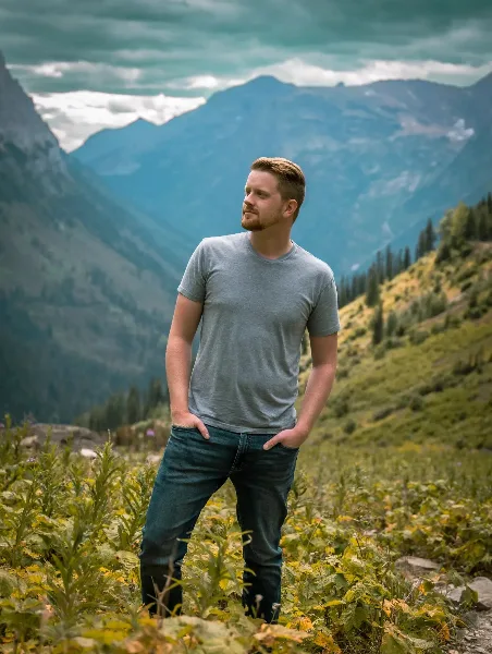 A man standing in a mountain meadow with a scenic backdrop of peaks and trees.