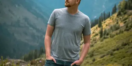 A man standing in a mountain meadow with a scenic backdrop of peaks and trees.