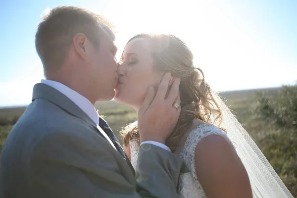 A bride and groom sharing a kiss outdoors with sunlight in the background.