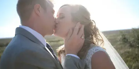 A bride and groom sharing a kiss outdoors with sunlight in the background.