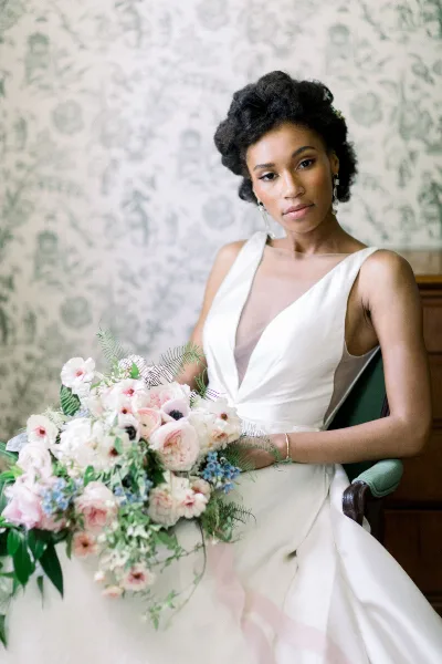 A bride in a white wedding dress holding a large floral bouquet while seated in a chair.