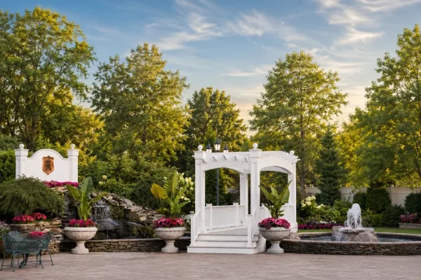 Outdoor wedding venue featuring a white arched structure, a stone waterfall, and a paved patio surrounded by manicured gardens and trees.