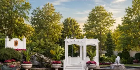 Outdoor wedding venue featuring a white arched structure, a stone waterfall, and a paved patio surrounded by manicured gardens and trees.