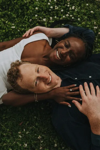 Candid photograph of a smiling interracial couple lying together in a grassy field with white flowers.