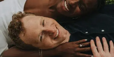 Candid photograph of a smiling interracial couple lying together in a grassy field with white flowers.