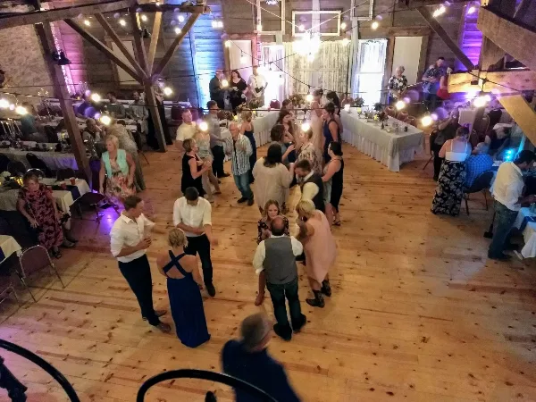 Guests dancing on a wooden floor inside a rustic barn venue with string lights and long dining tables.