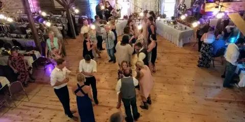 Guests dancing on a wooden floor inside a rustic barn venue with string lights and long dining tables.