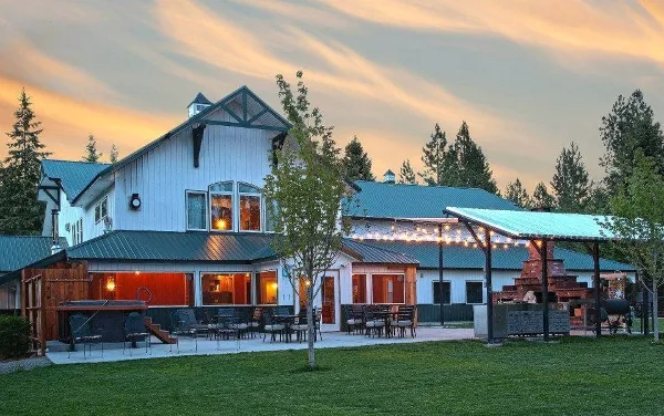 Exterior view of a white lodge with green metal roofs, outdoor patio seating, string lights, and surrounding trees at dusk.