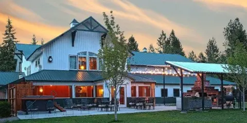 Exterior view of a white lodge with green metal roofs, outdoor patio seating, string lights, and surrounding trees at dusk.
