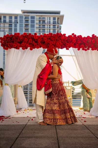 A bride and groom in traditional Indian wedding attire share a kiss under a red rose mandap during a wedding ceremony.