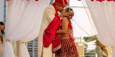 A bride and groom in traditional Indian wedding attire share a kiss under a red rose mandap during a wedding ceremony.