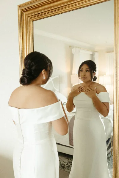 A bride in an off-the-shoulder white wedding dress with a styled updo looking at her reflection in a gold-framed mirror.