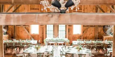 A rustic wooden event hall featuring multiple round tables set with white table linens, china, and glassware, ready for a wedding or event.