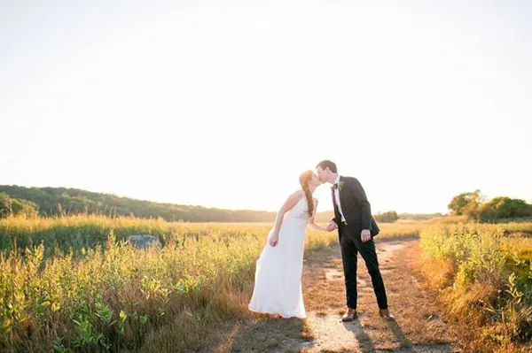 A bride and groom kissing on a dirt path in a field at sunset.