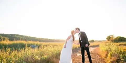 A bride and groom kissing on a dirt path in a field at sunset.