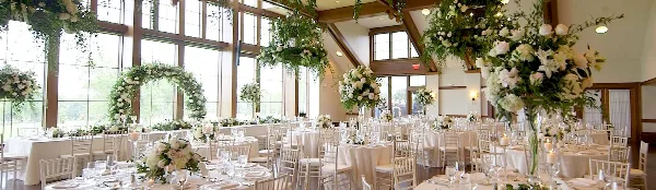 A spacious wedding reception hall featuring large windows, white tablecloths, and elaborate floral centerpieces and hanging arrangements.