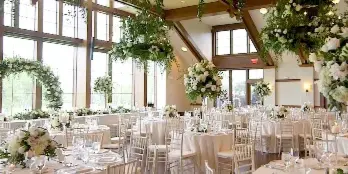A spacious wedding reception hall featuring large windows, white tablecloths, and elaborate floral centerpieces and hanging arrangements.
