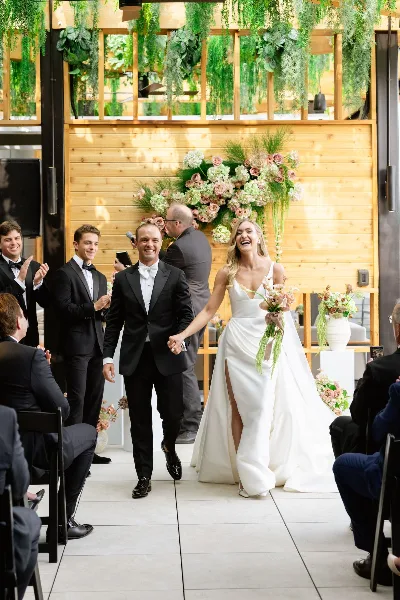 A bride and groom walking down the aisle in front of a large floral arrangement on a wooden wall.