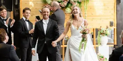 A bride and groom walking down the aisle in front of a large floral arrangement on a wooden wall.