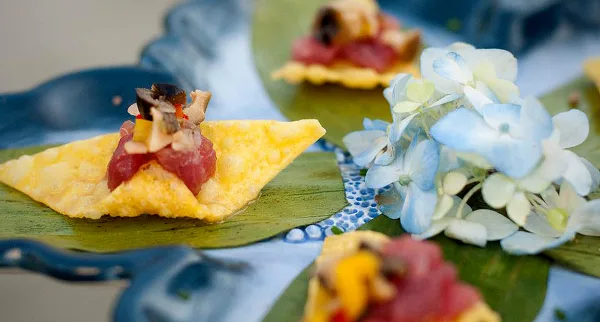 A plated appetizer featuring tuna tartare on a crisp, garnished with herbs and served on a blue ceramic plate with a small floral arrangement.