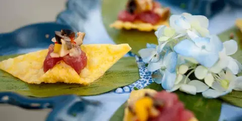 A plated appetizer featuring tuna tartare on a crisp, garnished with herbs and served on a blue ceramic plate with a small floral arrangement.