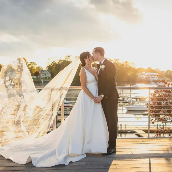 A bride and groom kissing on a wooden deck overlooking a marina with boats and a river at sunset.