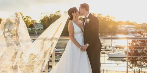 A bride and groom kissing on a wooden deck overlooking a marina with boats and a river at sunset.