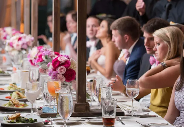 A long banquet table set with floral centerpieces and glassware, with a bride and groom seated among guests at a wedding reception.