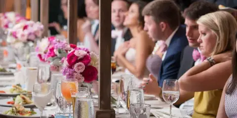 A long banquet table set with floral centerpieces and glassware, with a bride and groom seated among guests at a wedding reception.