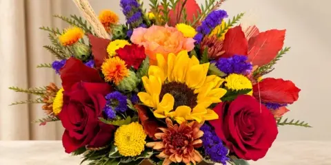 A mixed floral arrangement with sunflowers, red roses, and colorful mums in a bronze bowl vase.