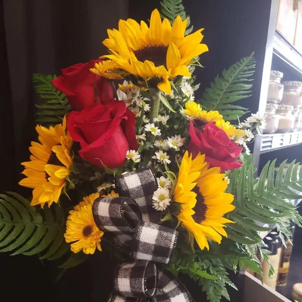 A floral arrangement featuring bright yellow sunflowers and red roses accented with green ferns and a black-and-white plaid ribbon.